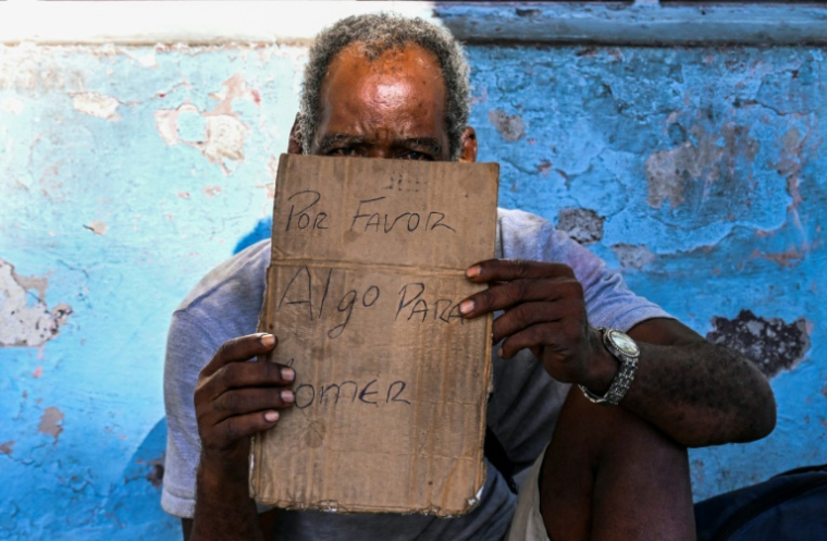 Un homme tient un morceau de carton sur lequel est écrit "S'il vous plaît, pour manger", alors qu'il mendie dans une rue de La Havane, le 21 juillet 2025 à Cuba ( AFP / Yamil LAGE )