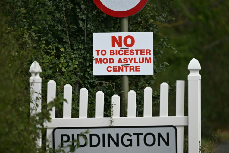 Un panneau indiquant l'entrée du village britannique de Piddington à côté d'un autre sur lequel on peut lire "Non au centre d’accueil pour demandeurs d’asile du ministère de la Défense à Bicester", le 15 septembre 2026 ( AFP / JUSTIN TALLIS )