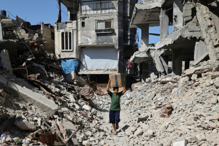 Un déplacé palestinien transportant un carton d'aide humanitaire marche au milieu de destructions à Nousseirat dans le centre de la bande de Gaza, le 19 octobre 2025 ( AFP / Eyad BABA )