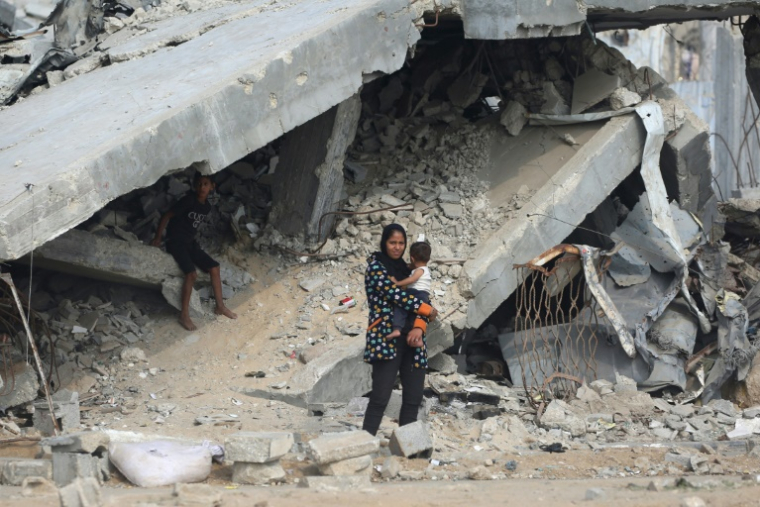 Une Palestinienne porte son enfant devant des bâtiments détruits le long d'une route près du camp de réfugiés de Nuseirat, dans le centre de la bande de Gaza, le 9 novembre 2025 ( AFP / Eyad Baba )