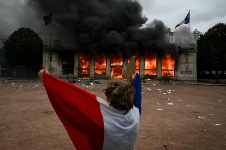 Un protestataire enveloppé dans un drapeau français devant l'antenne de l'Office du tourisme de Lyon en feu, lors d'une manifestation contre la vie chère, le 17 octobre 2026 ( AFP / Jeff PACHOUD )