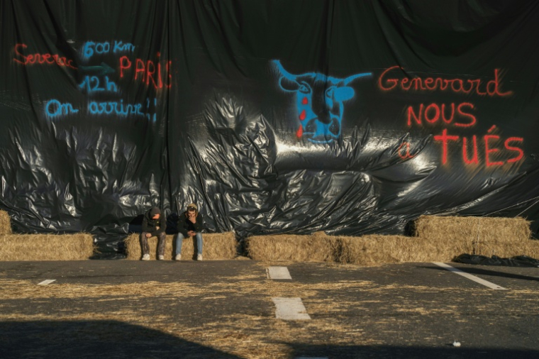 Des agriculteurs assis devant une bâche sur laquelle est écrit "Genevard nous a tués" (d) lors du blocage de l'autoroute A75 à Séverac-d'Aveyron, le 14 décembre 2025 dans l'Aveyron ( AFP / Idriss Bigou-Gilles )