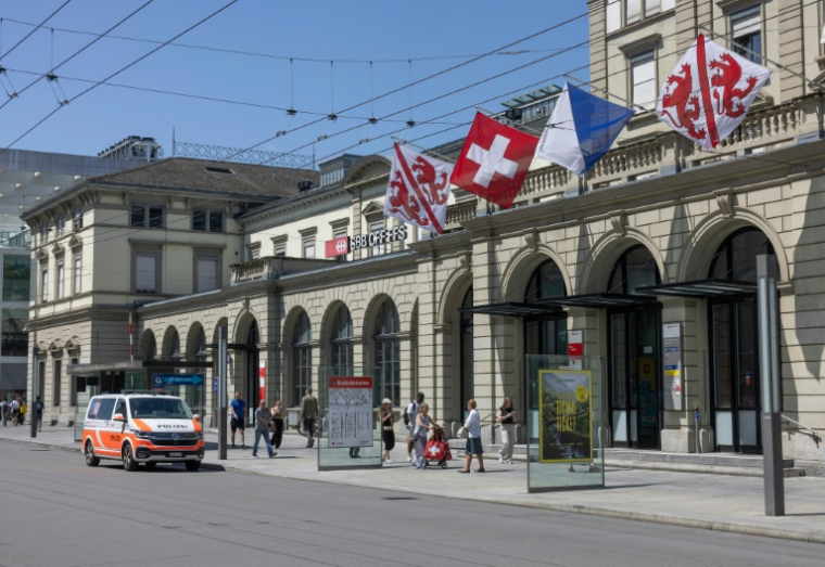 Des passants défilent devant un véhicule de police stationné devant la gare centrale de Winterthur, près de Zurich, où un homme a blessé trois personnes à l'arme blanche le 28 mai 2026 ( AFP / ARND WIEGMANN )