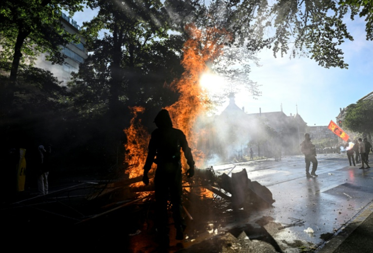 Manifestant devant une poubelle en feu lors d'une manifestation anti-G7 à Genève, le 14 juin 2026 ( AFP / Gabriel Monnet )