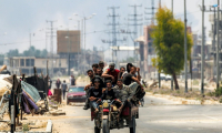 Des hommes à l'arrière d'un tricycle le long de la route de Salaheddin à Deir al-Balah, dans le centre de la bande de Gaza, le 21 juillet 2025, tandis que derrière eux s'élèvent des volutes de fumée provenant des bombardements israéliens ( AFP / Eyad BABA )