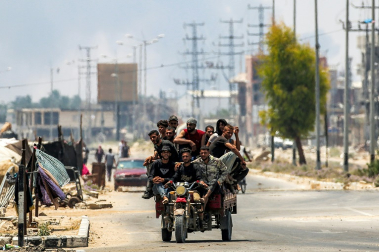 Des hommes à l'arrière d'un tricycle le long de la route de Salaheddin à Deir al-Balah, dans le centre de la bande de Gaza, le 21 juillet 2025, tandis que derrière eux s'élèvent des volutes de fumée provenant des bombardements israéliens ( AFP / Eyad BABA )